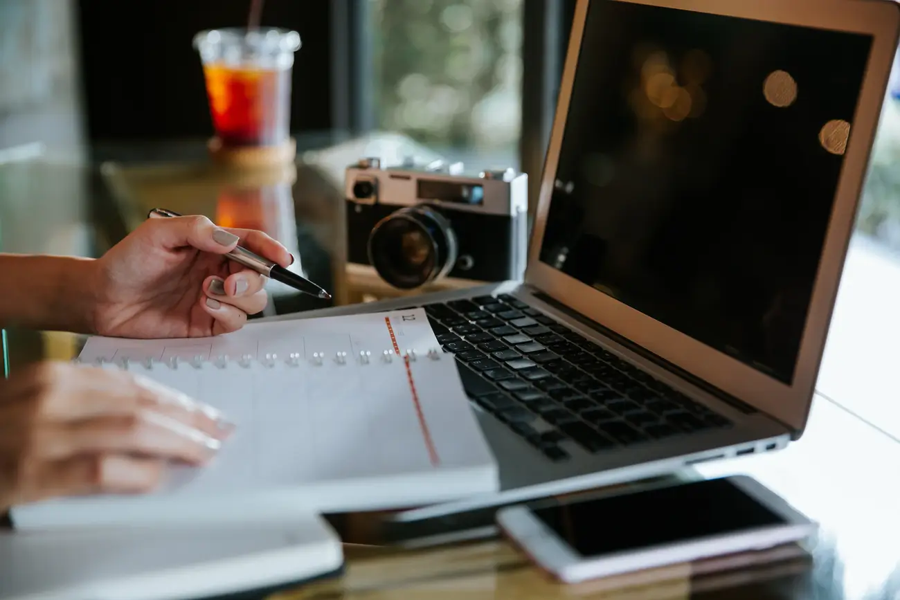 Person writing in notebook beside laptop with camera, drink, and smartphone
