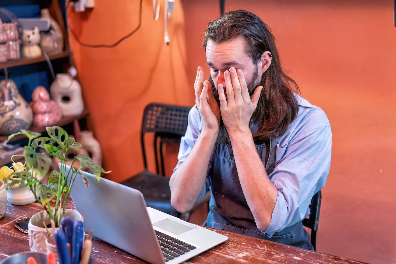 Person in apron at table with laptop, covering face in stress