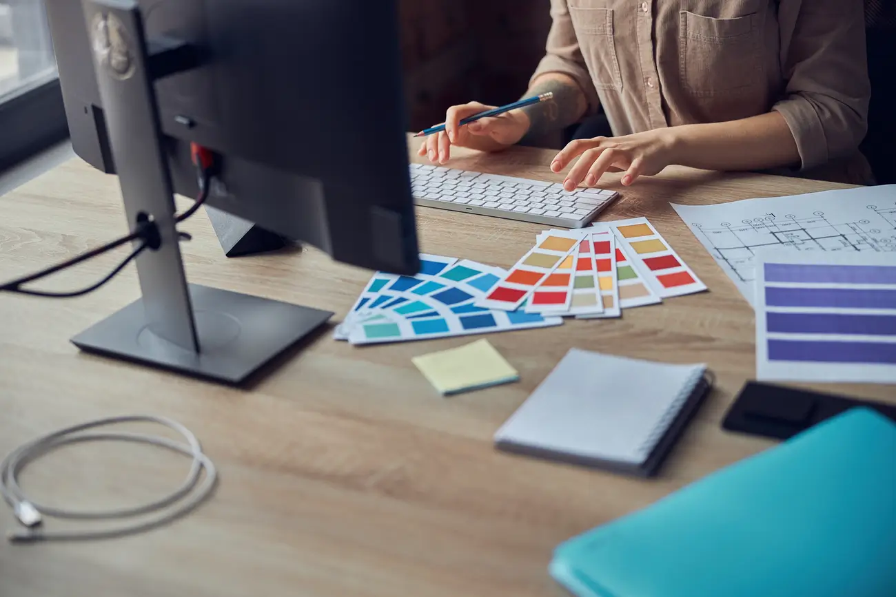 Person at desk with computer, swatches, papers, notebook, sticky notes, and folder
