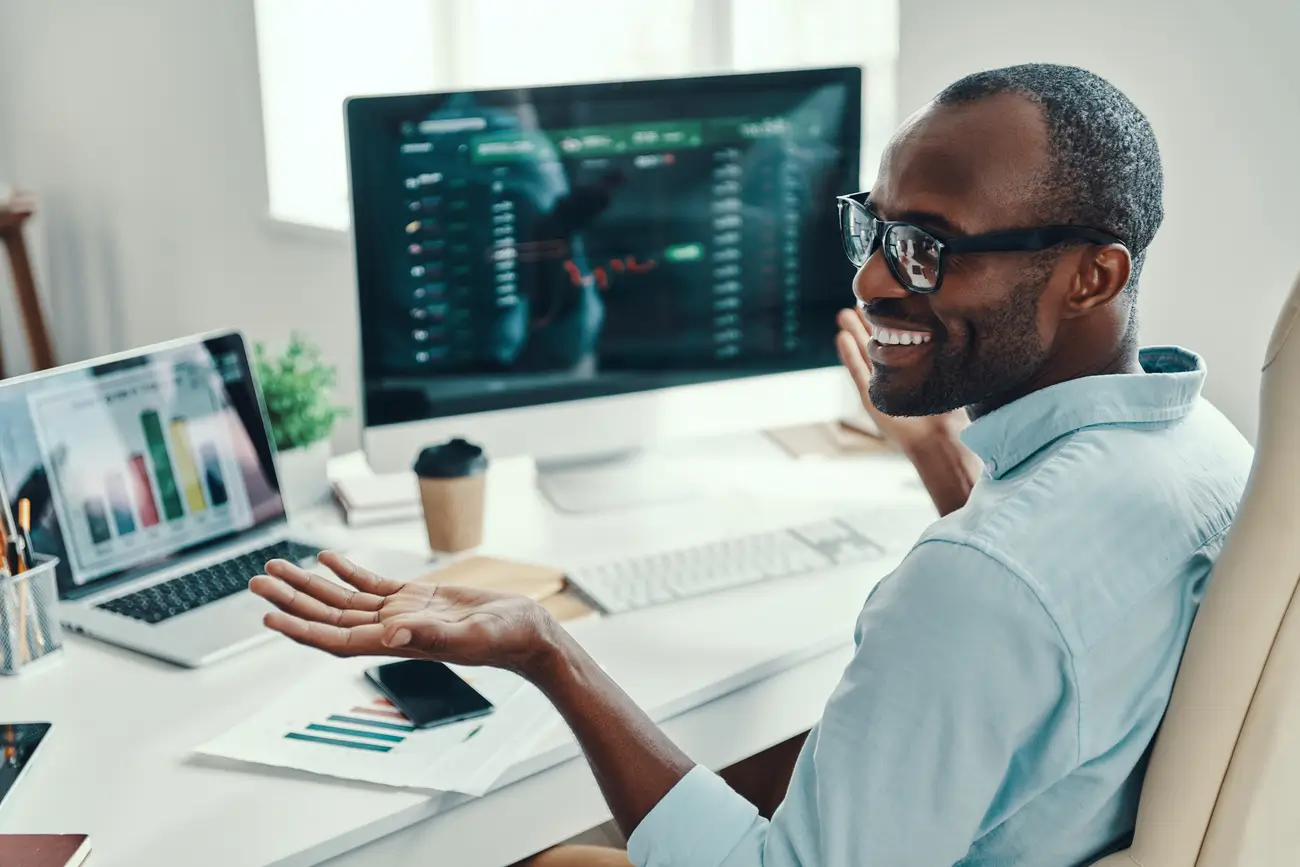 Man with glasses at desk with graphs on screens, smiling and gesturing