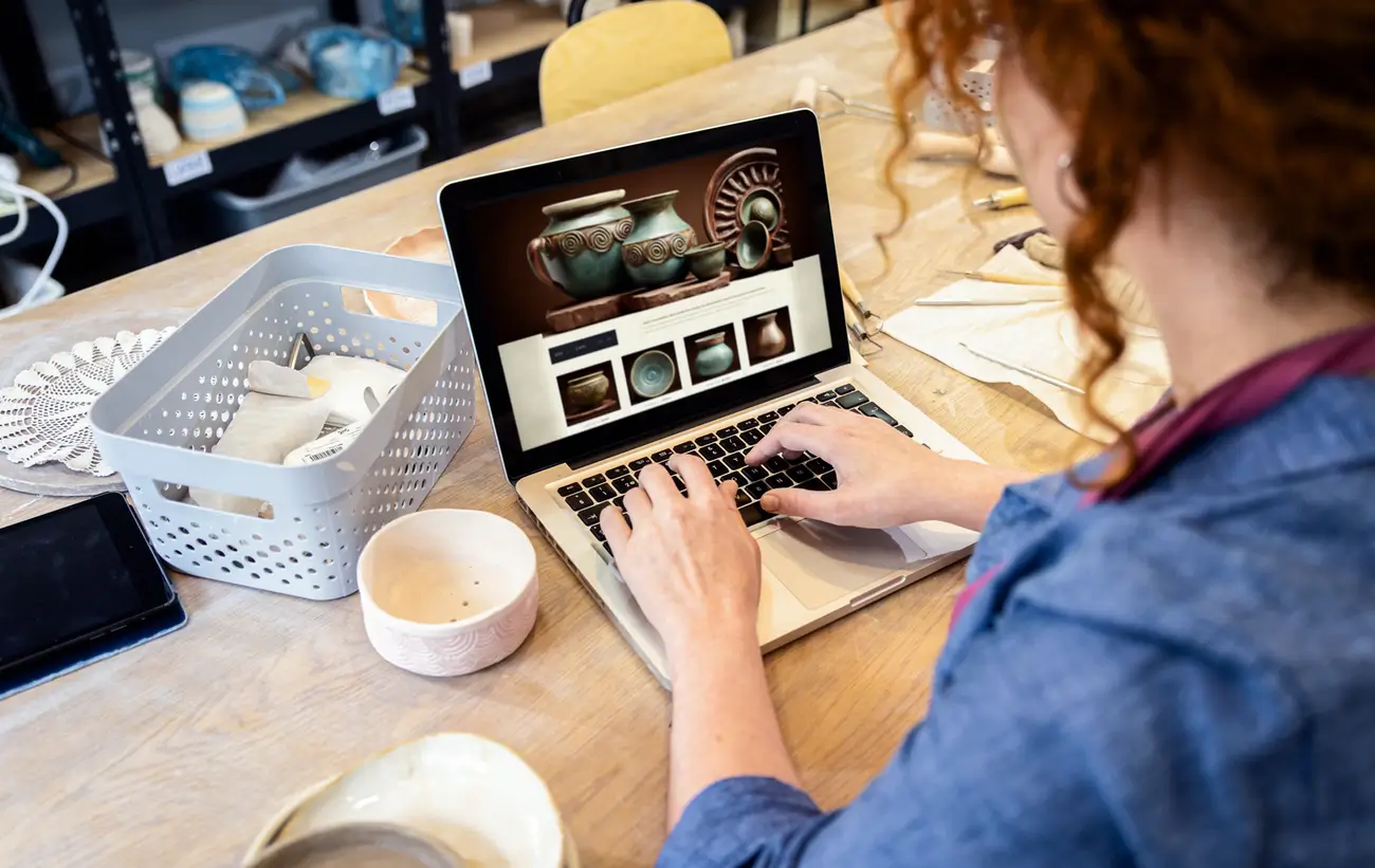 Person using laptop to browse pottery website at table with ceramics and tools