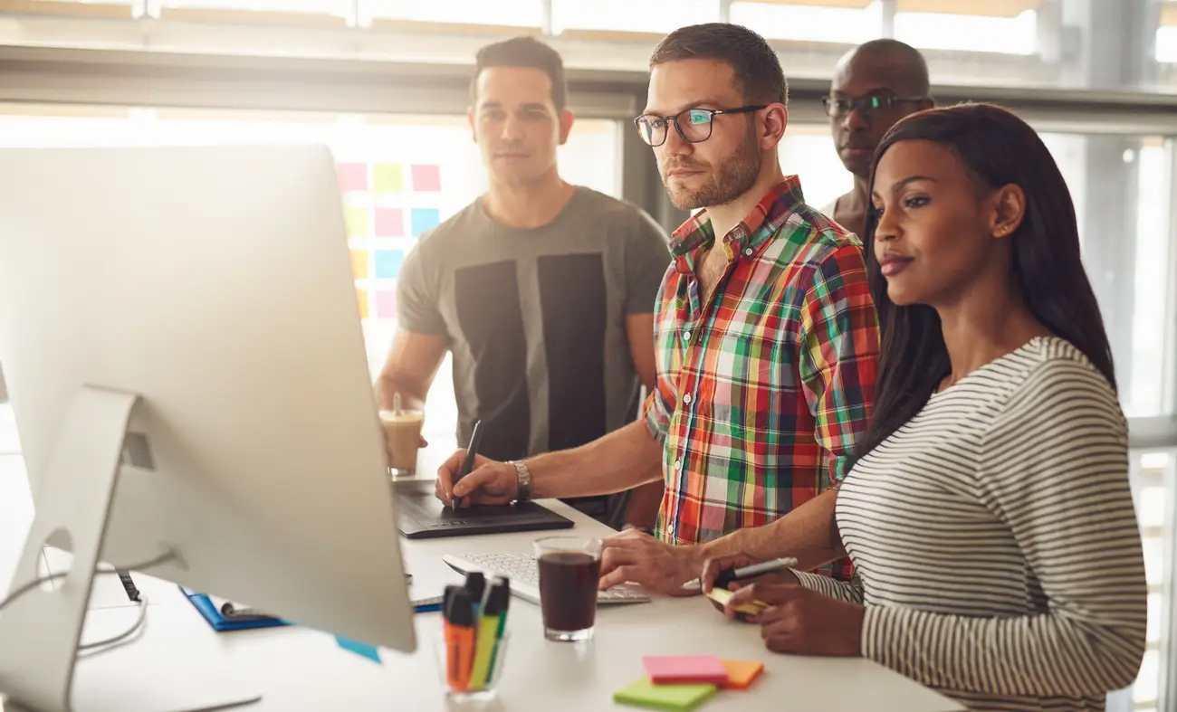 Four people stand at a desk in a modern office, looking at a large monitor while one uses a mouse and stylus.