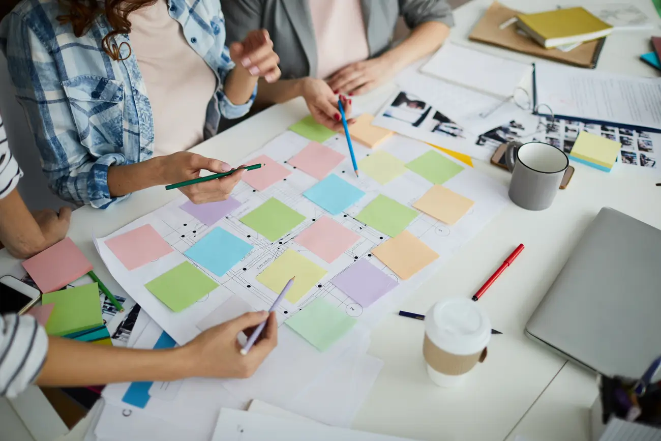 Three people collaborate at a table with colorful sticky notes, documents, and drinks, planning a project.