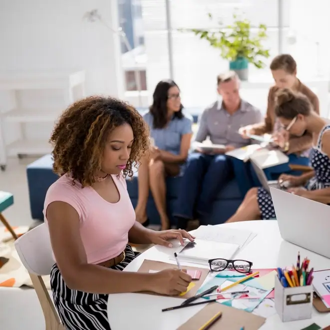 Woman working at a desk while colleagues meet in the background.