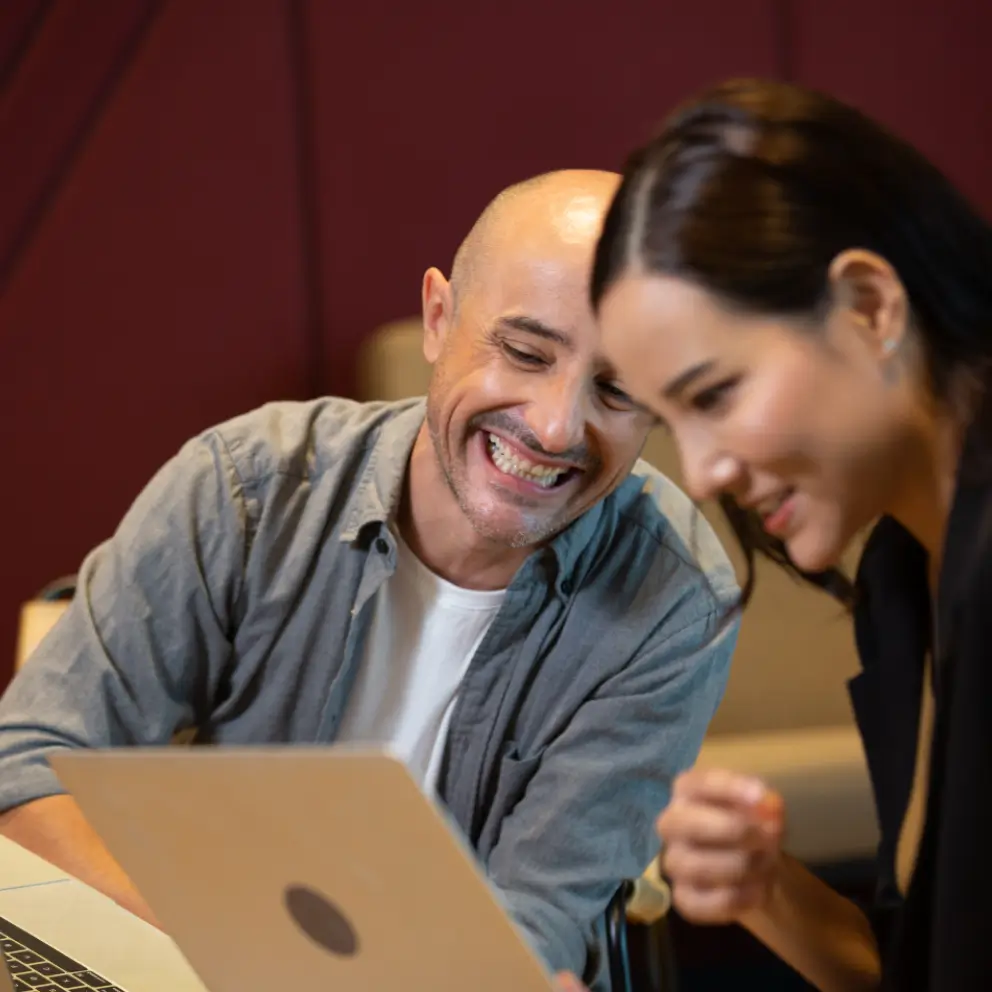 A man and a woman sit side by side, smiling and looking at a laptop screen together in an indoor setting.