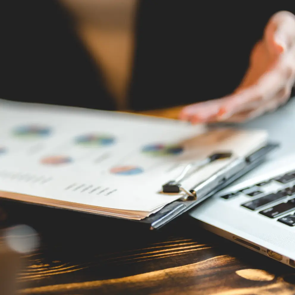 A person gestures toward a clipboard with printed charts and graphs next to an open laptop on a wooden desk.