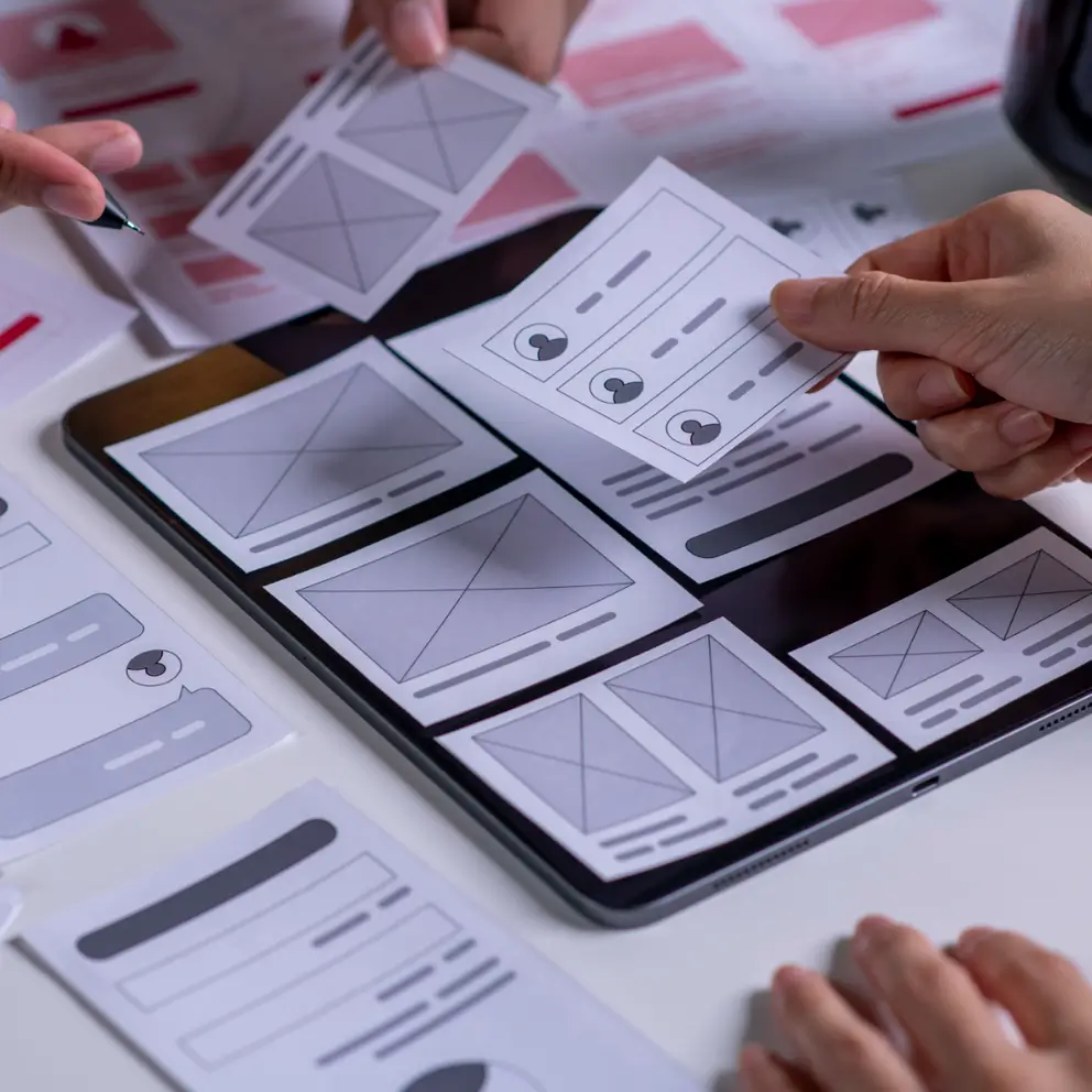 Hands arranging paper wireframes on a tablet, planning a website or app user interface with various layout sketches and design elements.